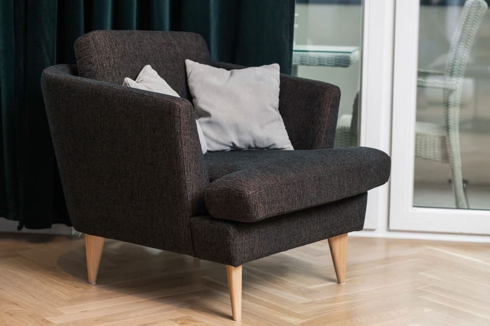 A modern armchair with textured upholstery, paired with a soft throw pillow. Positioned by the window, it's ideal for relaxing with a book.