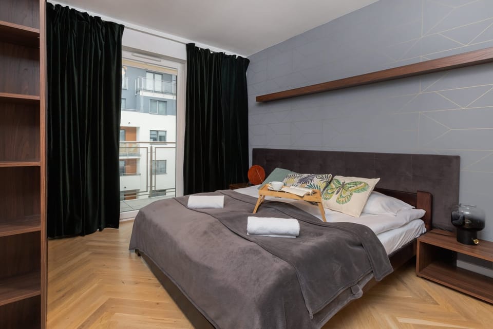 A cozy bedroom with a double bed, dark curtains, and a view of the cityscape through a wide window. The neutral decor creates a calming atmosphere.