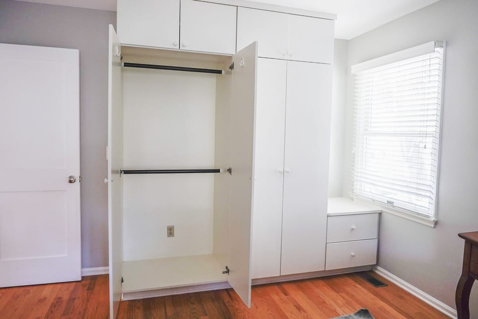 Open white wardrobe with multiple compartments next to a window with blinds in a room with wooden flooring.