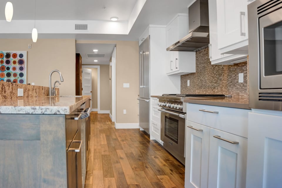 Open concept kitchen with white cabinets and island with granite counter tops in front, and stainless steel appliances.