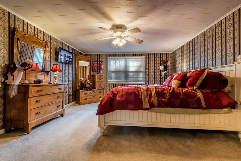 Bedroom with patterned wallpaper, white wood bed dressed in red and gold linens, and wooden dressers with mirrors.