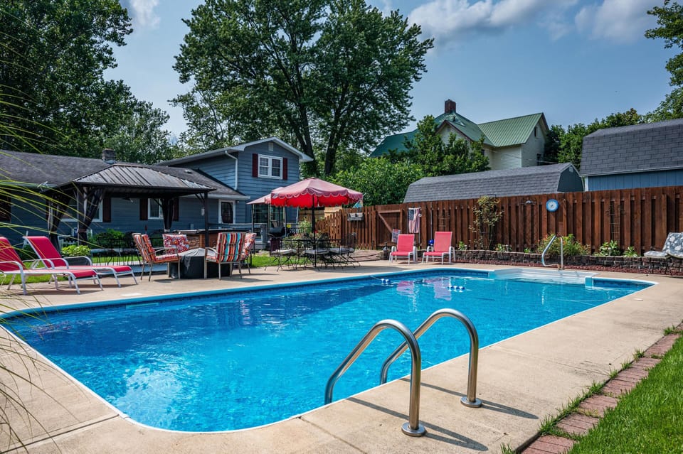 Private backyard with rectangular pool, red loungers, umbrella-shaded table, and blue-gray house exterior.