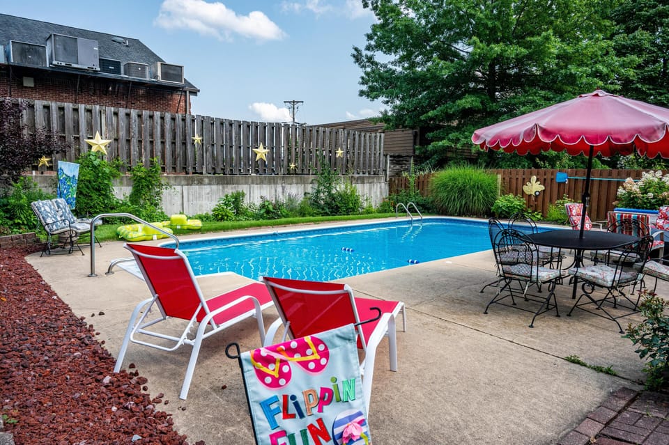 Rectangular pool surrounded by concrete deck, red loungers, black metal table with umbrella, and garden greenery.