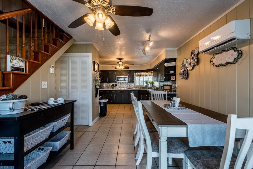 Dining area with white wooden chairs and table runner, adjacent to tile floor kitchen and wood staircase.