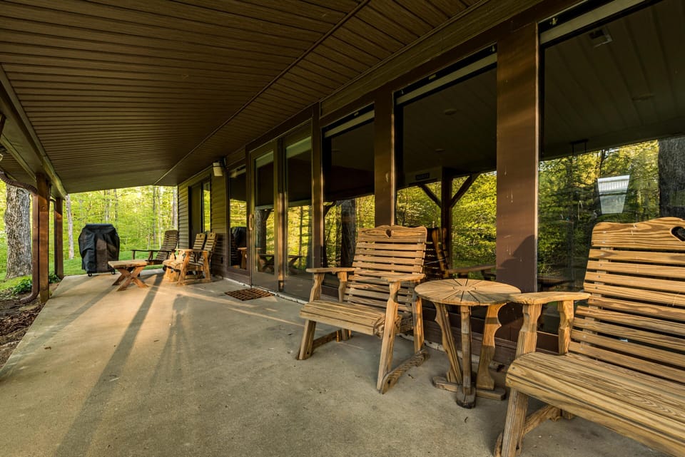 Covered patio with wood slat chairs, small matching tables, and a charcoal grill overlooking green trees.