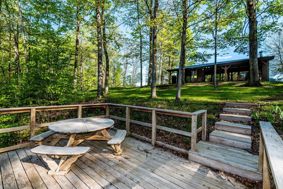 Wide wooden deck with round picnic table and benches, stairs leading up to cabin through green lawn and trees.