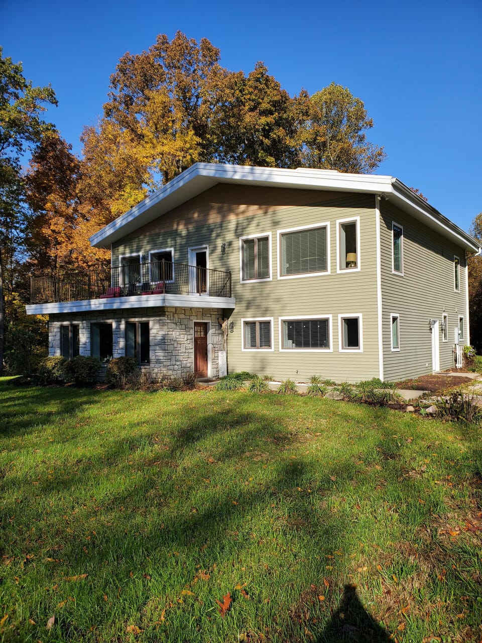 Two-story house with olive-green siding, stone accents, and a balcony overlooking a grassy yard.