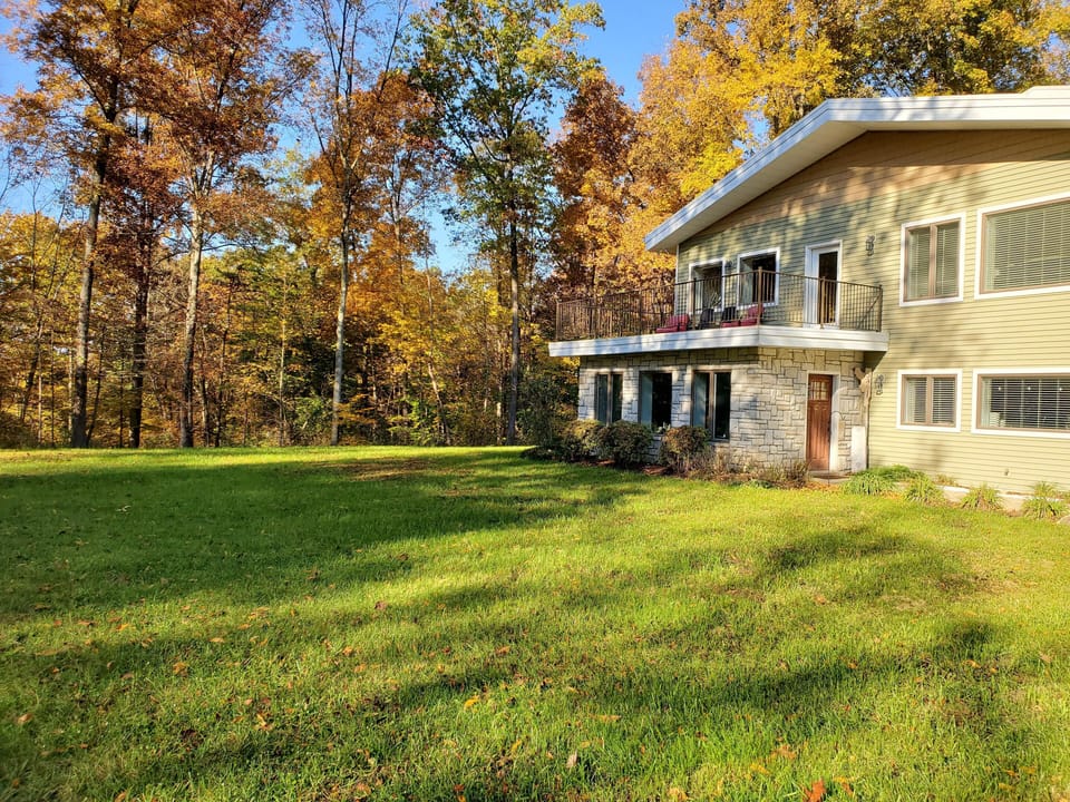 Grassy backyard bordered by tall trees with autumn foliage beside a two-story house featuring stone and beige siding.