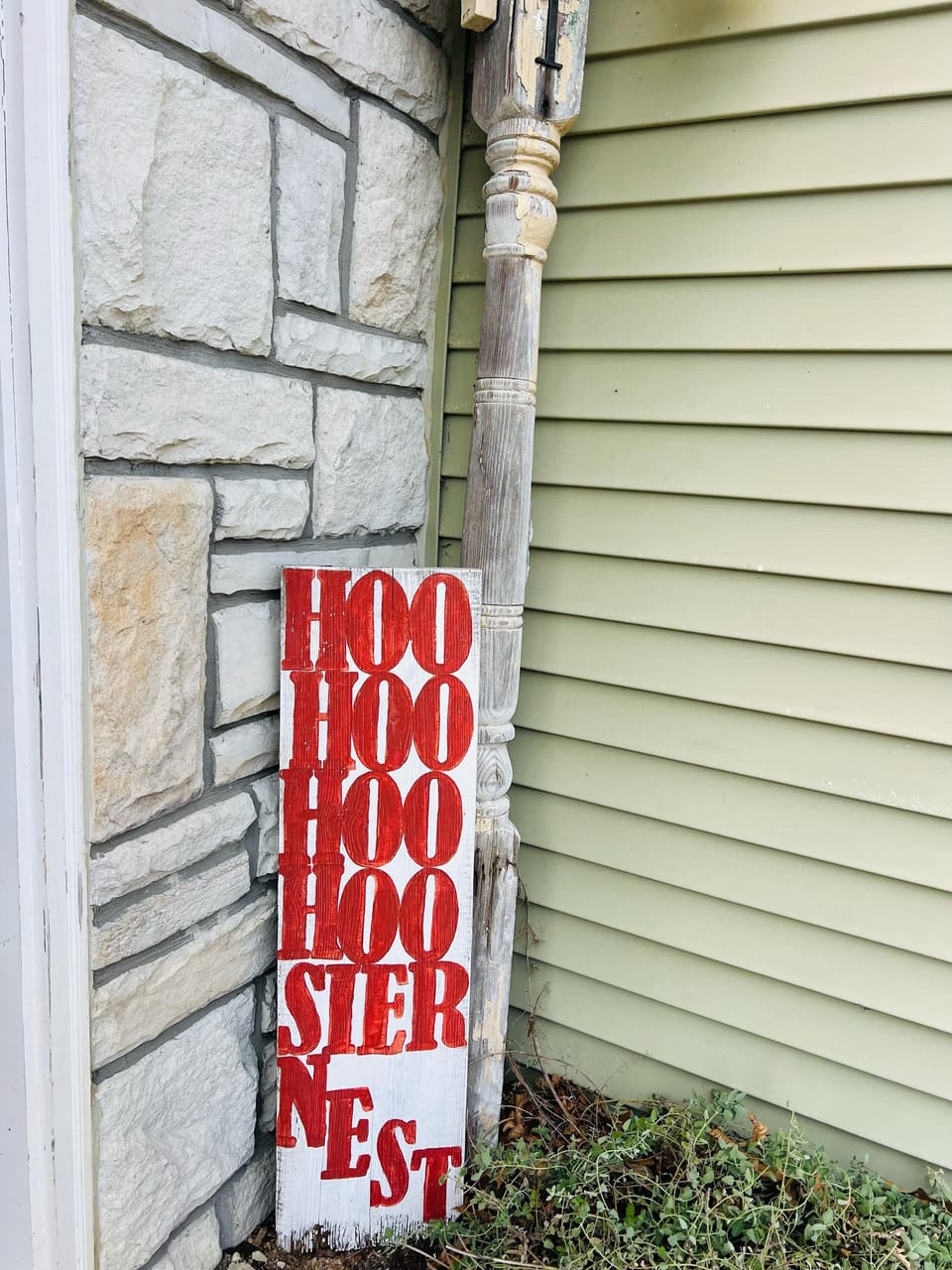 Corner of the house featuring light green vinyl siding, a stone wall, and a white wooden sign with red letters.