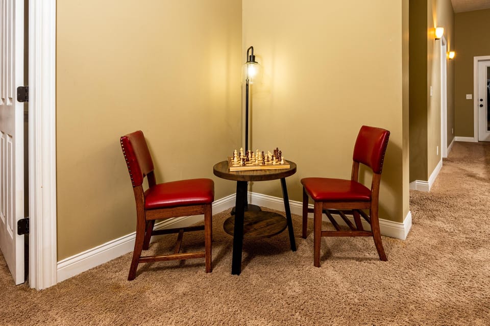Corner nook with a round wooden table holding a chess set, flanked by two red leather-upholstered chairs.