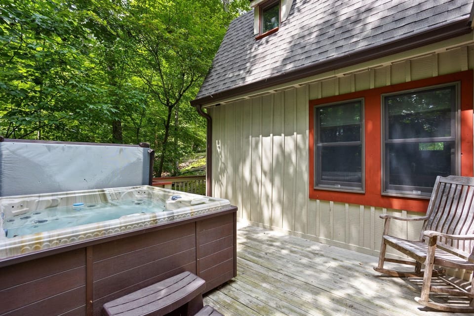 Wood deck with a brown hot tub, matching steps, weathered rocking chair, and two red-trimmed windows.