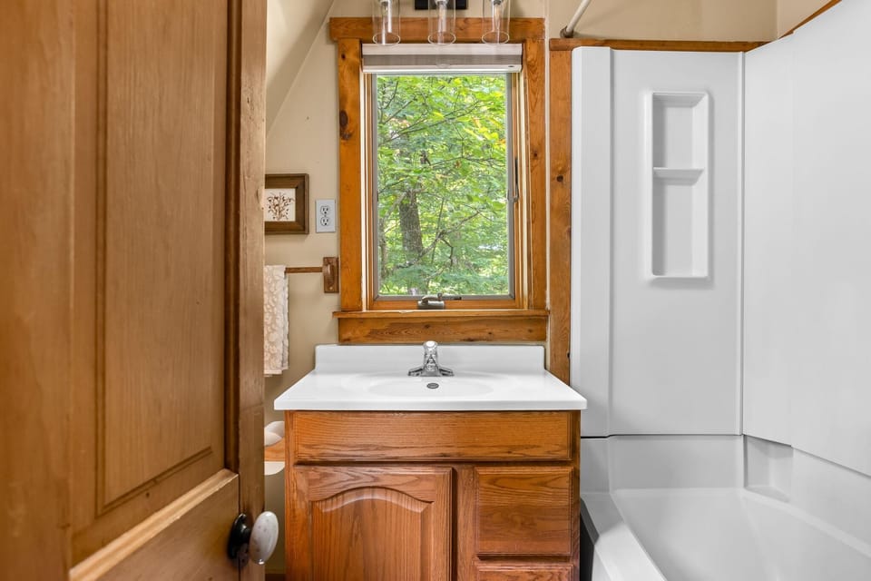 Bathroom features a white sink atop wood cabinetry, a white tub with built-in shelves, and a window framed by natural wood.