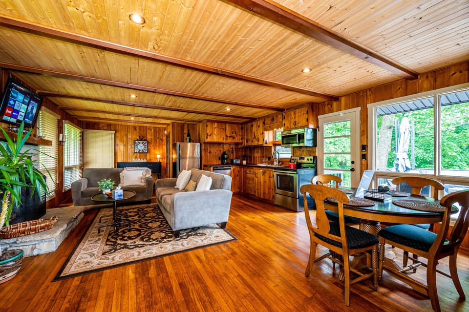 Open-concept living area with knotty pine walls, stone fireplace, stainless steel kitchen, and dining table by large windows.
