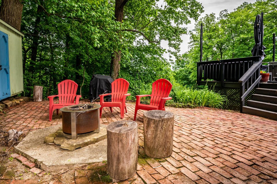 Outdoor fire pit area with red Adirondack chairs, rustic log stools, brick patio, and surrounding green trees.