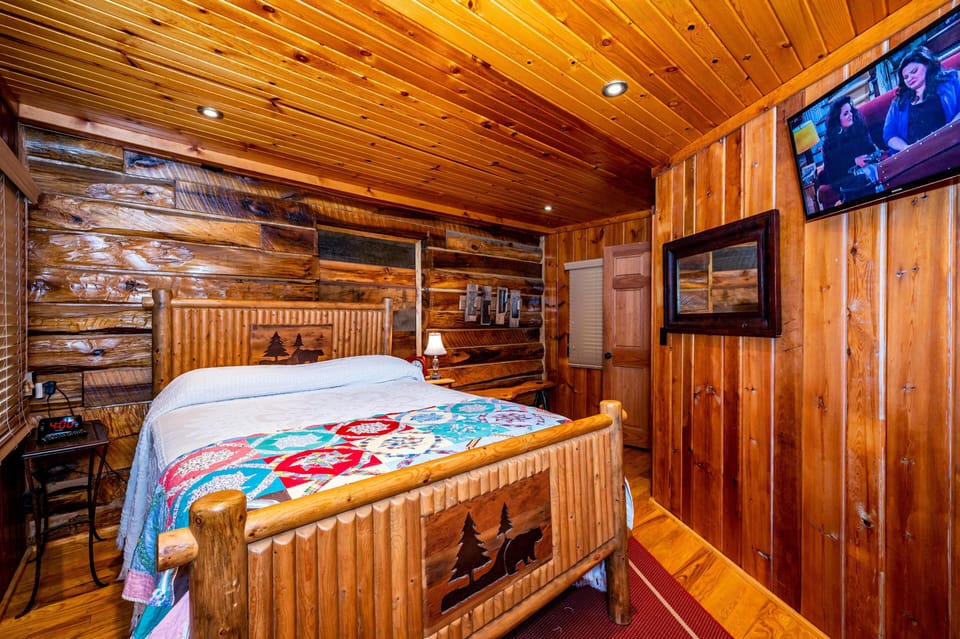 Rustic bedroom with log-framed bed featuring carved bear and tree motifs, colorful quilt, and wood-paneled walls.