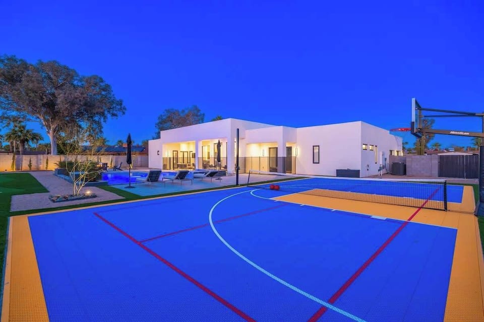 Modern desert villa showcases an expansive outdoor sports court with basketball hoop, heated pool, and sleek architecture glowing warmly under deep twilight skies.