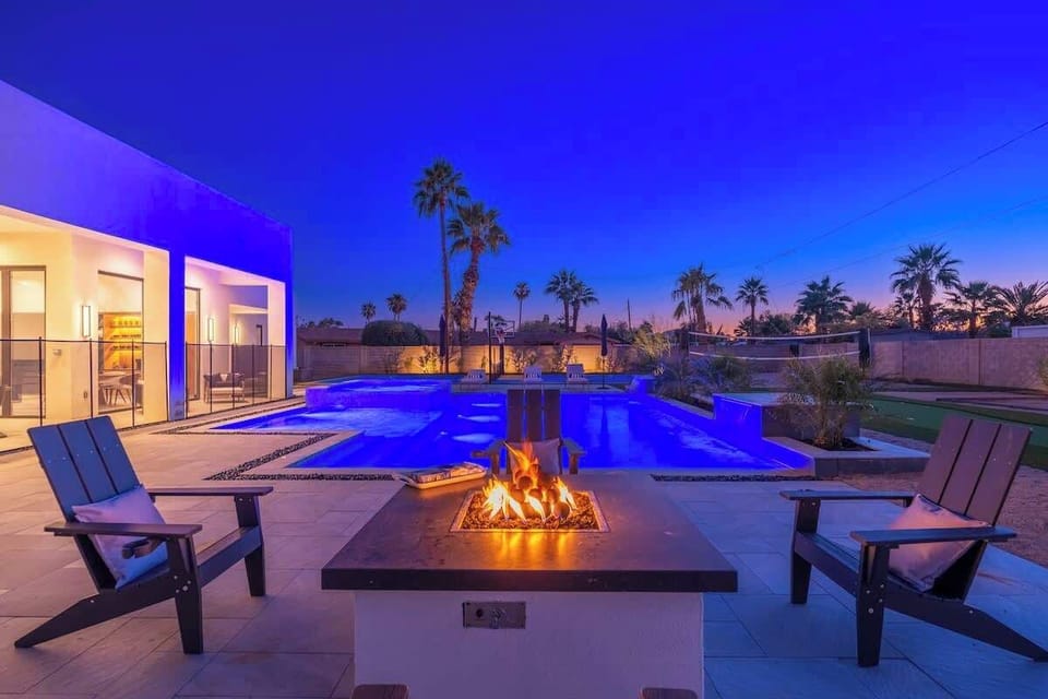 Desert twilight surrounds this modern retreat, featuring an outdoor fire pit, illuminated pool, and palm-studded landscape perfect for memorable evenings under the stars.