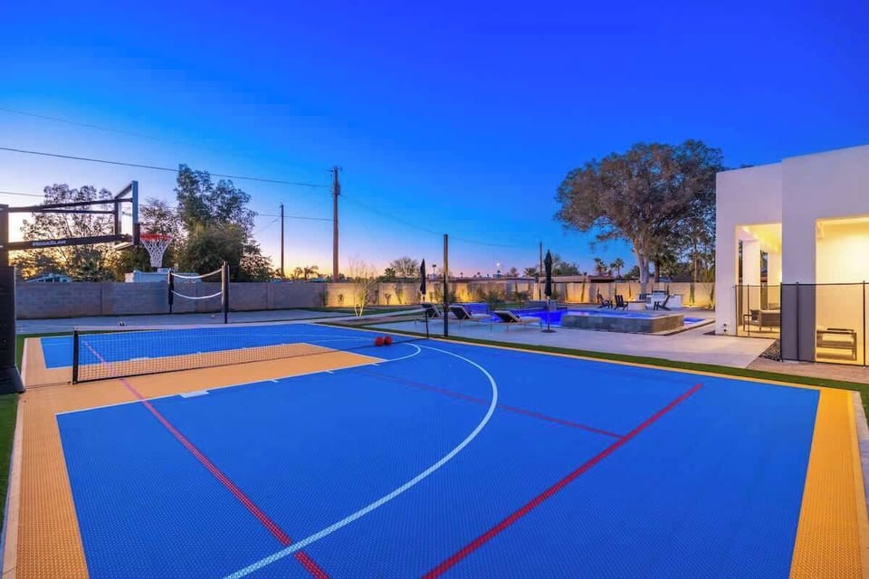 Full-size outdoor basketball court with volleyball setup awaits, backed by a glowing pool area perfect for post-game relaxation under desert skies.
