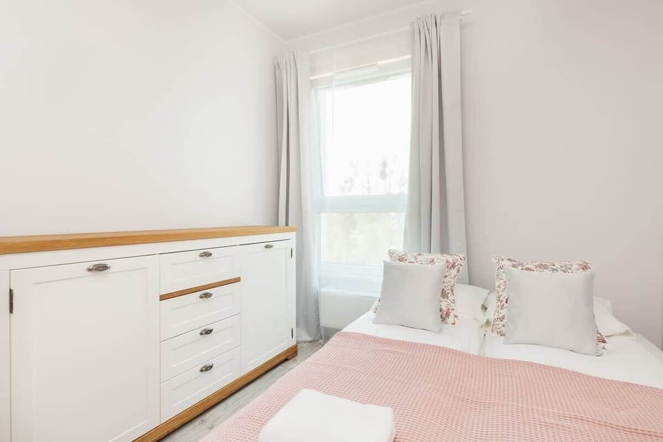 Another bedroom with a white bed, pink bedding, and a chest of drawers. The window is dressed with light curtains.