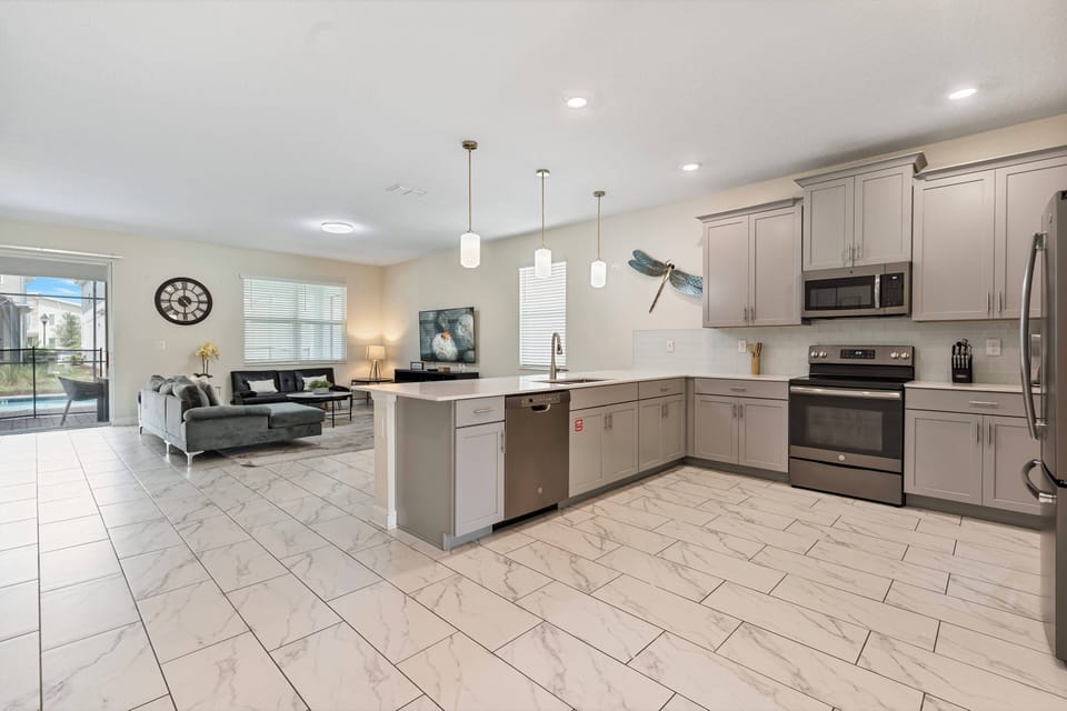 Discover a spacious kitchen that flows into a cozy living area, equipped with stainless steel appliances and stylish light fixtures. The soft gray cabinetry pairs beautifully with bright tile flooring for a warm, modern feel.
