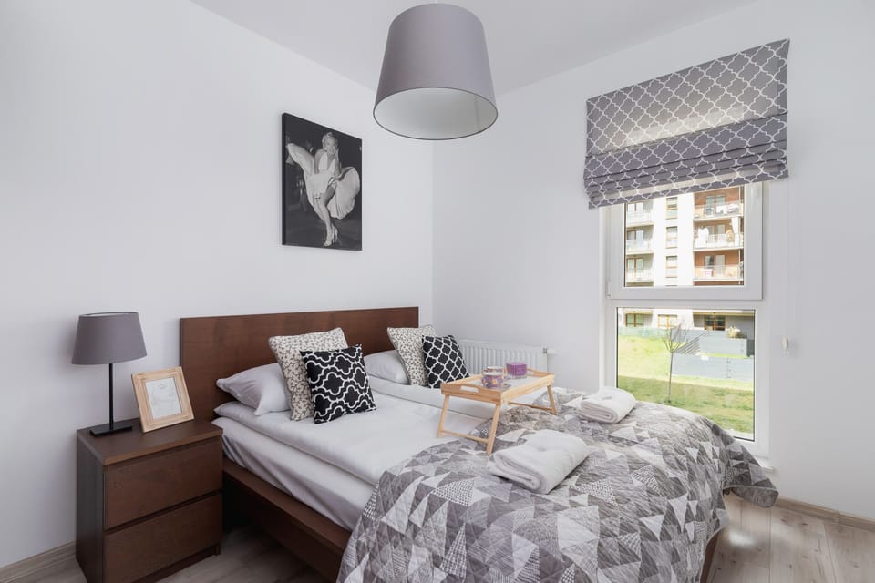 A stylish bedroom with a comfortable double bed, gray bedding, and a matching nightstand. A modern table lamp and a decorative framed artwork add to the cozy and elegant atmosphere.