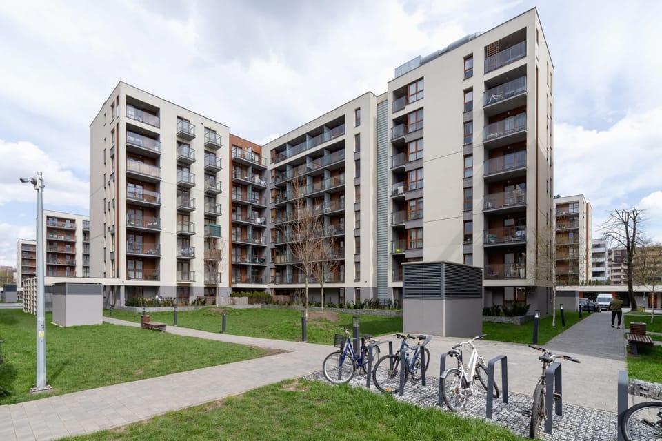 An exterior shot of a modern apartment complex with multiple balconies and a green courtyard. The building features a contemporary architectural design.