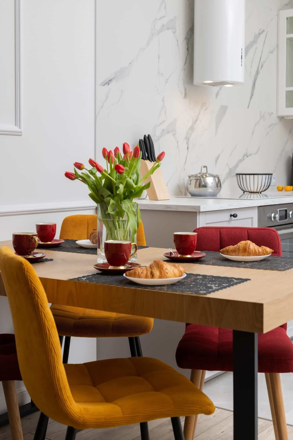 A close-up of the dining table set with vibrant red accents, flowers, and a welcoming breakfast arrangement.

