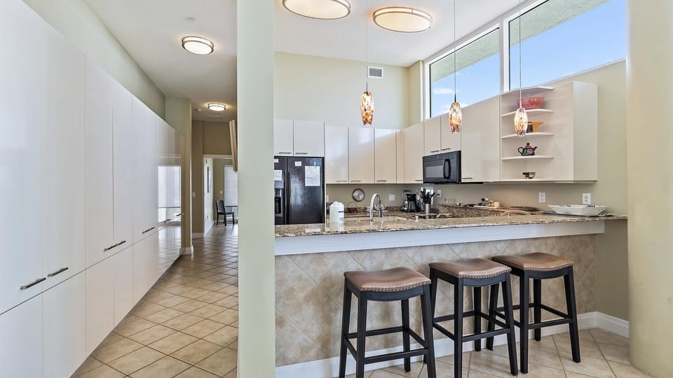 A modern kitchen with granite countertops, additional bar stool seating at island. Hanging pendant lights illuminate the counter area.