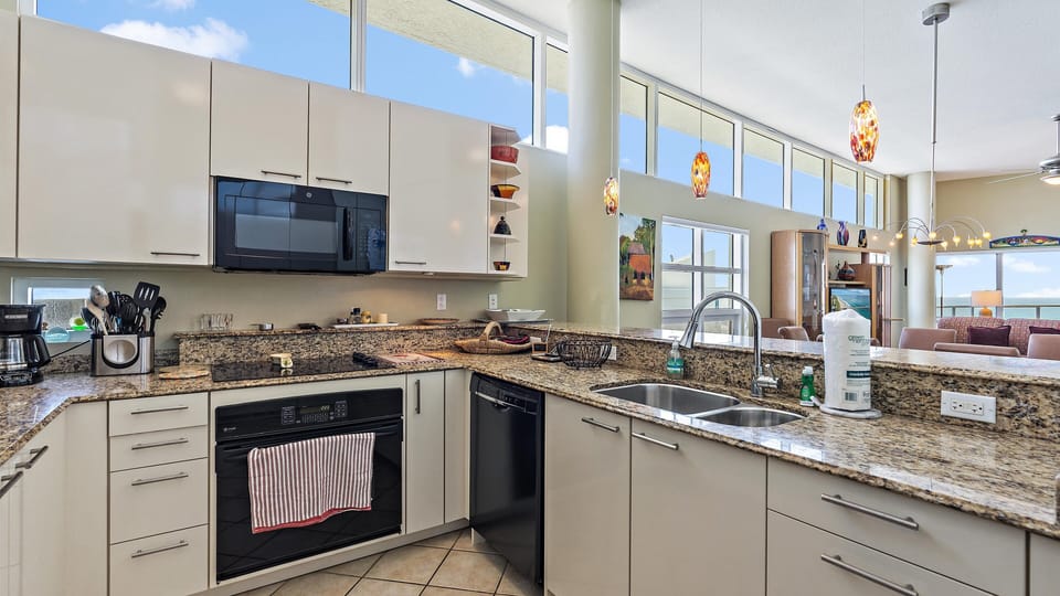 Modern kitchen with granite countertops, a double sink, and built-in appliances. Sunlit with large windows and pendant lights. Open to dining and main living space.