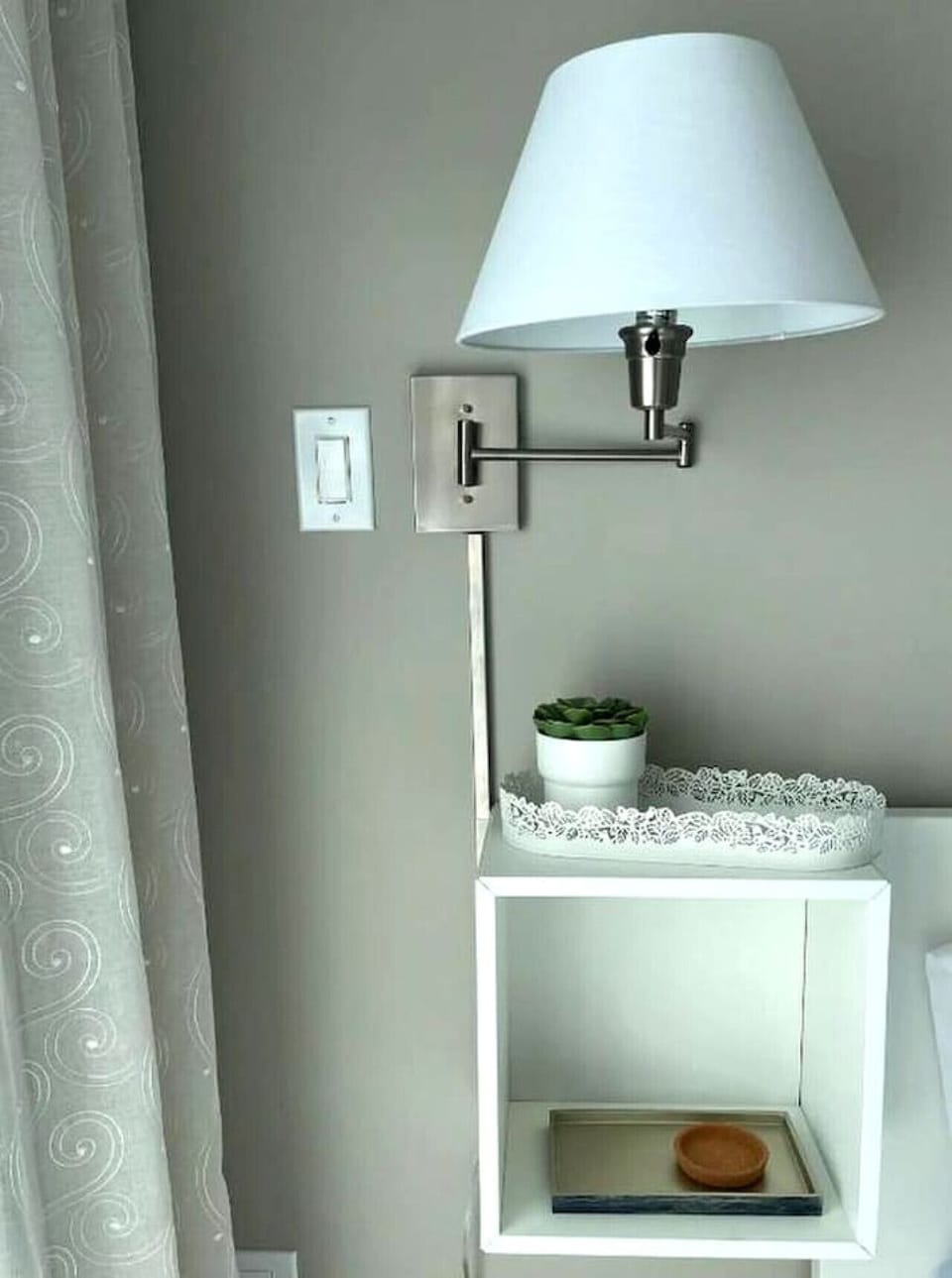 Modern wall-mounted lamp with a white shade above a minimalist white shelf featuring a small potted plant.