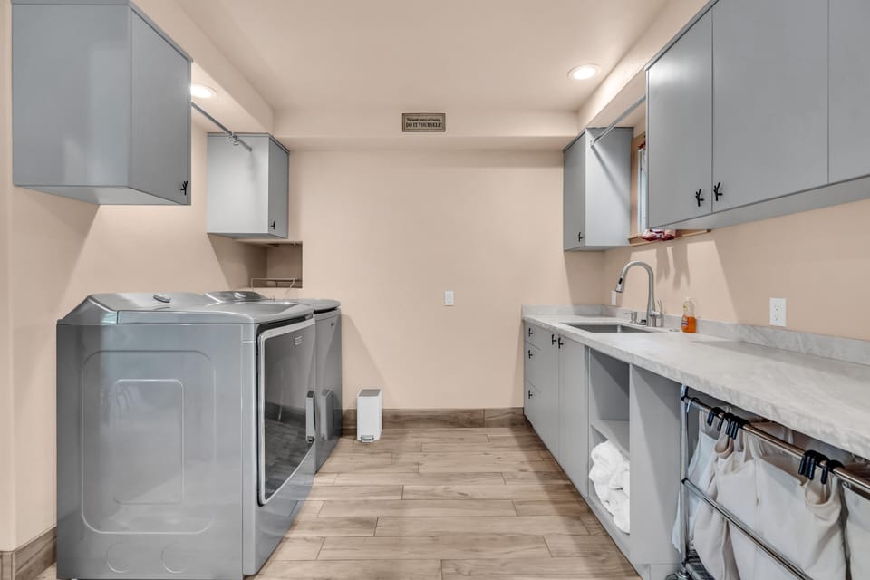 Laundry room with light gray cabinets, marble countertop, stainless steel washer and dryer, and wood-look tile floor.
