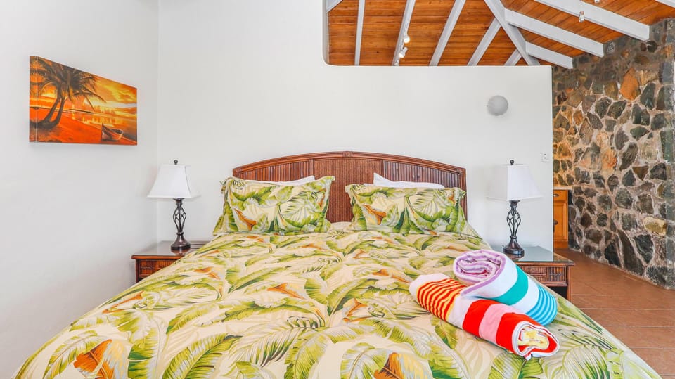 King bed with tropical leaf bedding sits beneath vaulted wood ceiling and beside a stone accent wall.