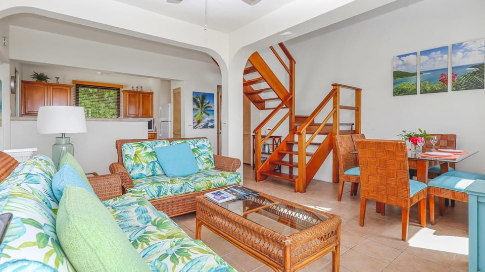 Living room with floral-patterned wicker sofas, glass-top coffee table, and wooden staircase rising beside the dining area.