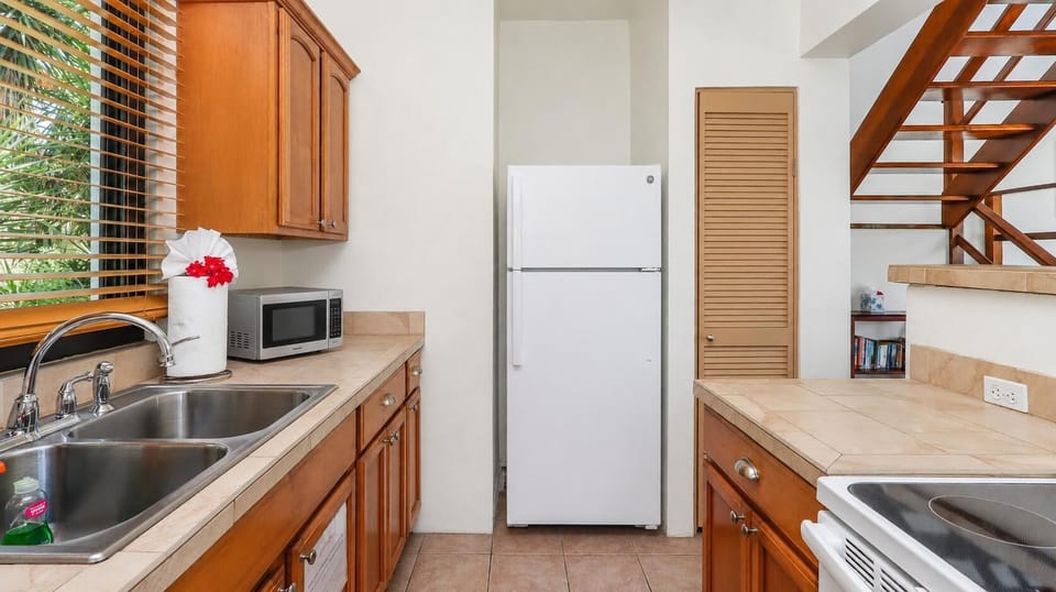 Tile countertops and wooden cabinets frame a white refrigerator and stainless steel double sink beneath wood-blind window.