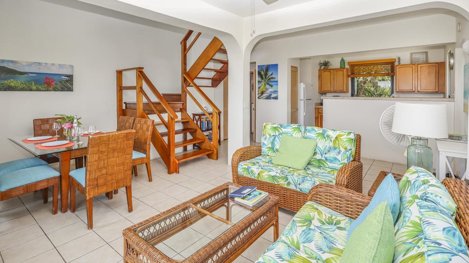 Living and dining area with wicker furniture, tropical cushions, glass-top table, and wooden staircase leading upstairs.