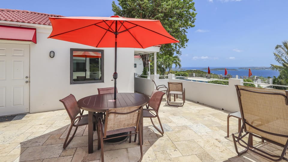 Outdoor terrace with tan stone tile flooring, brown metal dining set, bright red umbrella, and ocean views.
