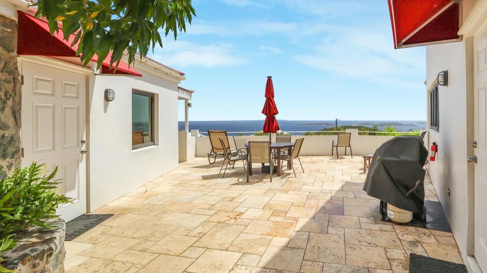 Sunlit stone patio with metal dining set, red umbrella, covered grill, and ocean views beyond white walls.
