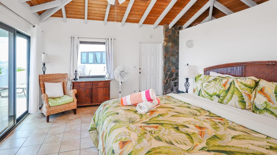 King bed with tropical leaf print bedding faces sliding glass doors and a wicker armchair on tile floors.