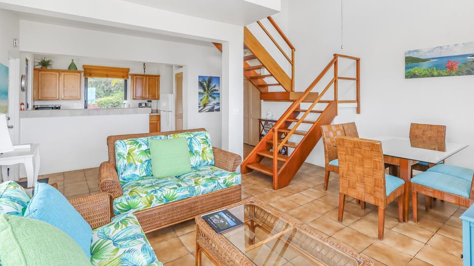 Wicker sofas with tropical-print cushions face a glass-top coffee table on tile floors beneath a wood staircase.