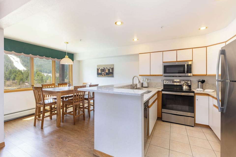 A kitchen and dining area with wooden furniture, stainless steel appliances, a large window with a green valance, and a view of a forested landscape. A painting hangs on the wall above the dining table.
