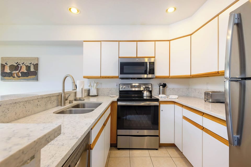 A modern kitchen with white cabinets, stainless steel appliances including a refrigerator, microwave, and oven, a double sink, and a marble countertop. A wall-mounted artwork is visible in the background.