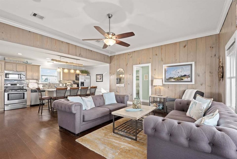Living room seating area with gray sofas, armchair, wood accent walls, table lamp, wall mirror, and coastal artwork.
