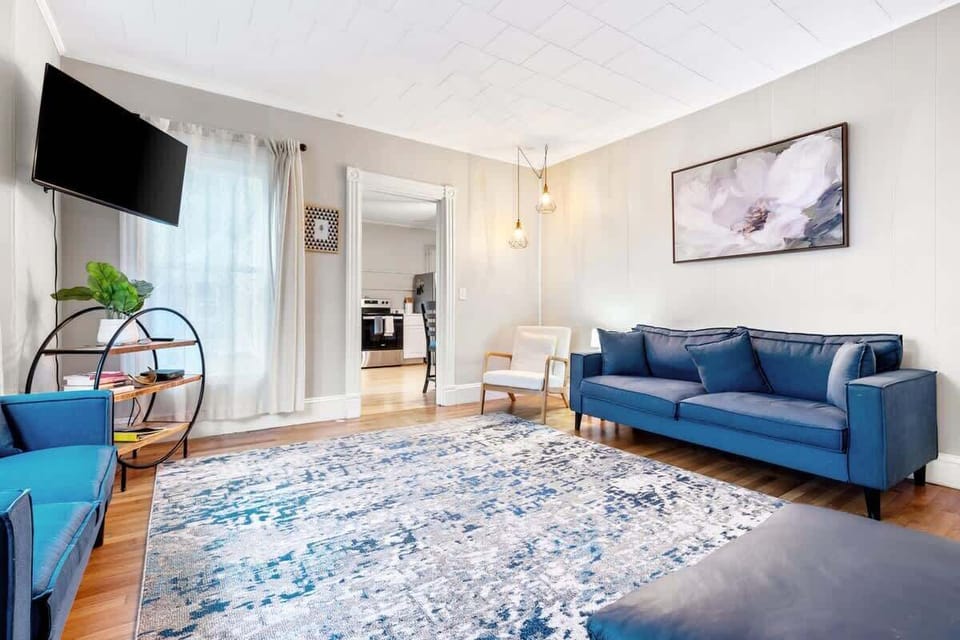 Large living room includes multiple seating options, a mounted TV, and a round shelf, with pendant lighting and an open view into the kitchen.