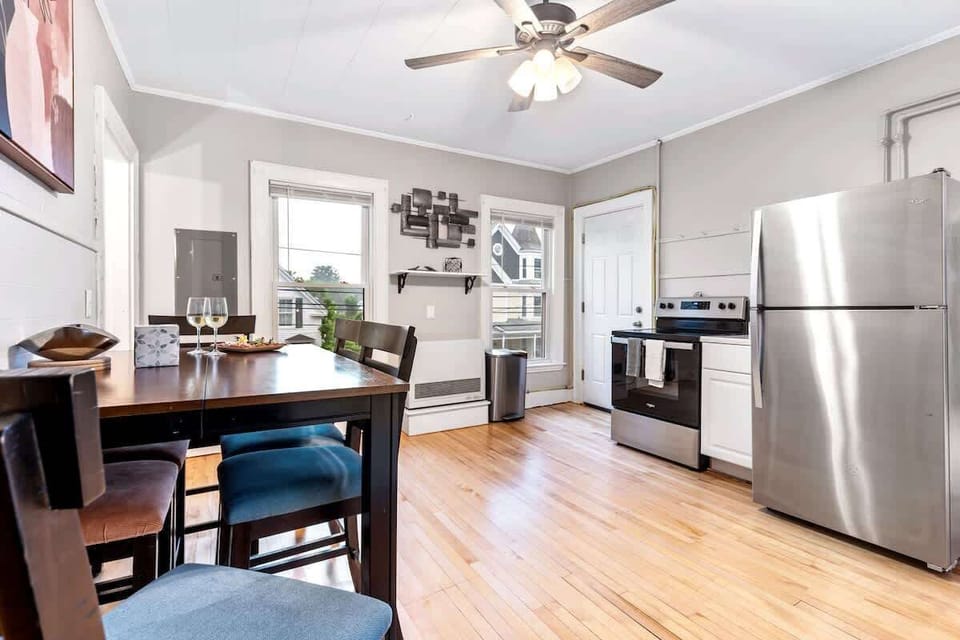The dining area sits by two large windows that fill the space with natural light, next to a modern kitchen with stainless steel appliances.