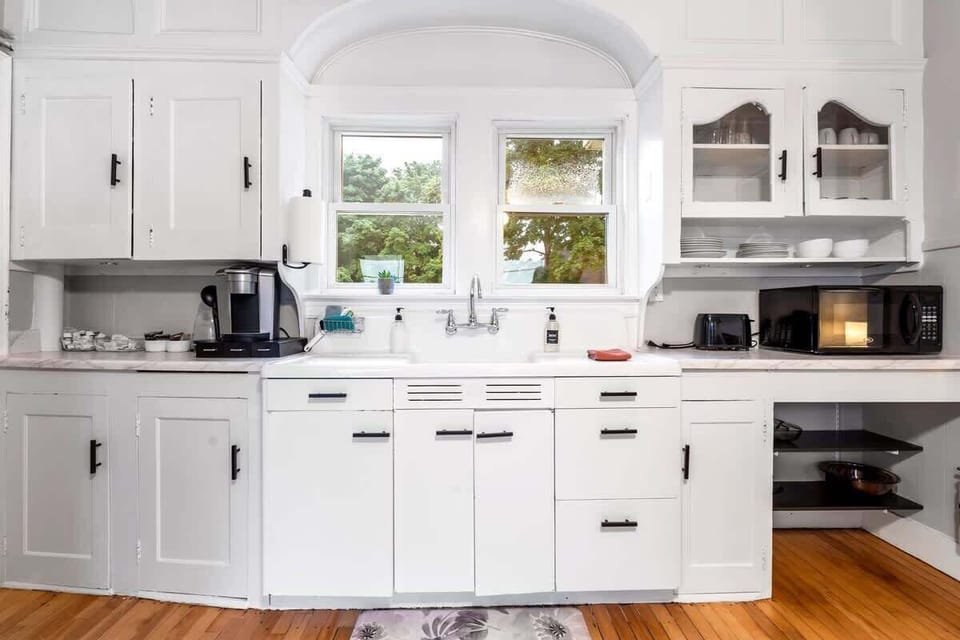 This kitchen features classic white cabinetry, a double window above the farmers sink, built-in shelving with dishes, and essentials like a microwave, toaster, and Keurig coffee maker.