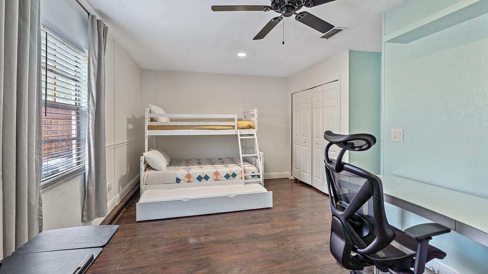 A bedroom with wooden floors features a bunk bed, a desk with a chair, a ceiling fan, and a window with closed curtains.