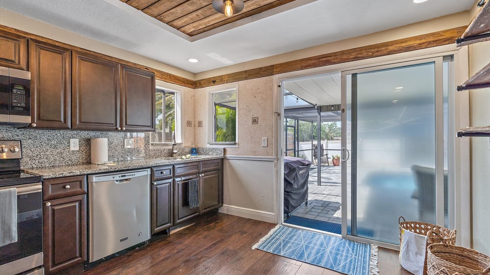 Modern kitchen with dark wood cabinets, granite countertops, stainless steel appliances, and hardwood flooring. A sliding glass door opens to a patio with outdoor seating and a barbecue grill.