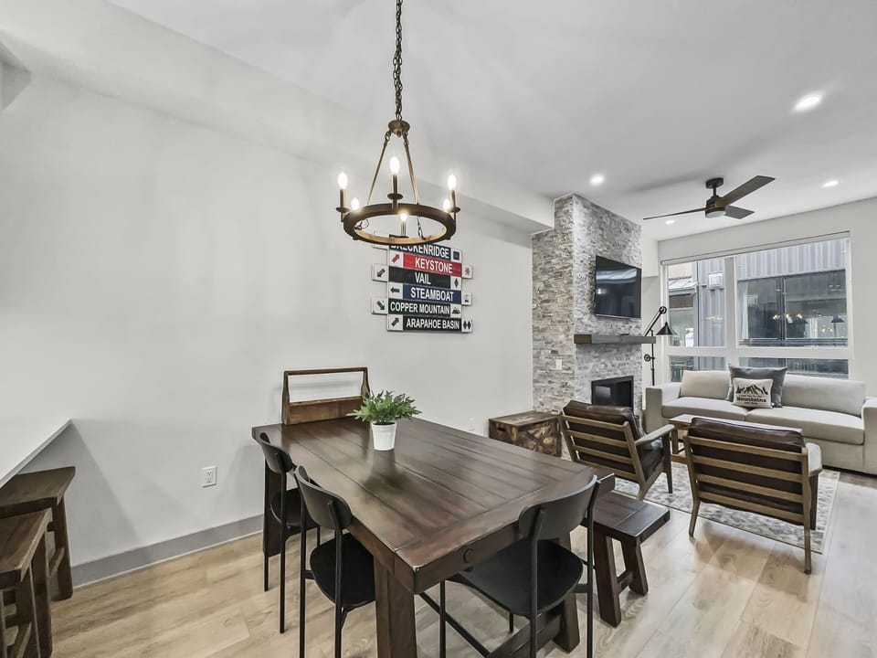 A modern living and dining area with a wooden dining table and chairs, a lounge area with a couch and wooden chairs, a stone fireplace, and a ceiling fan. A sign with multiple locations is on the wall.