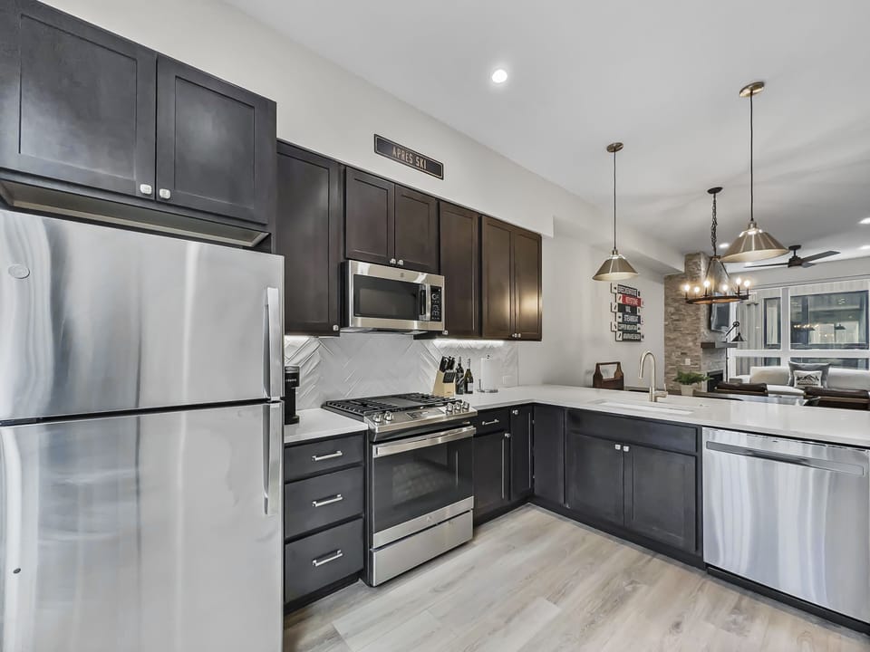 Modern kitchen with stainless steel appliances, dark cabinets, a gas stove, overhead microwave, and a dual sink. Wooden flooring and a view into the dining and living areas.
