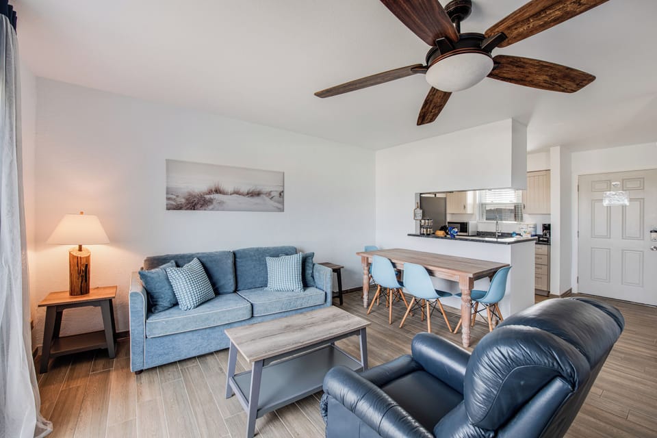 Relax in your coastal-styled living space with blue accents, wood-beam ceiling fan, and modern open layout connecting to the kitchen.